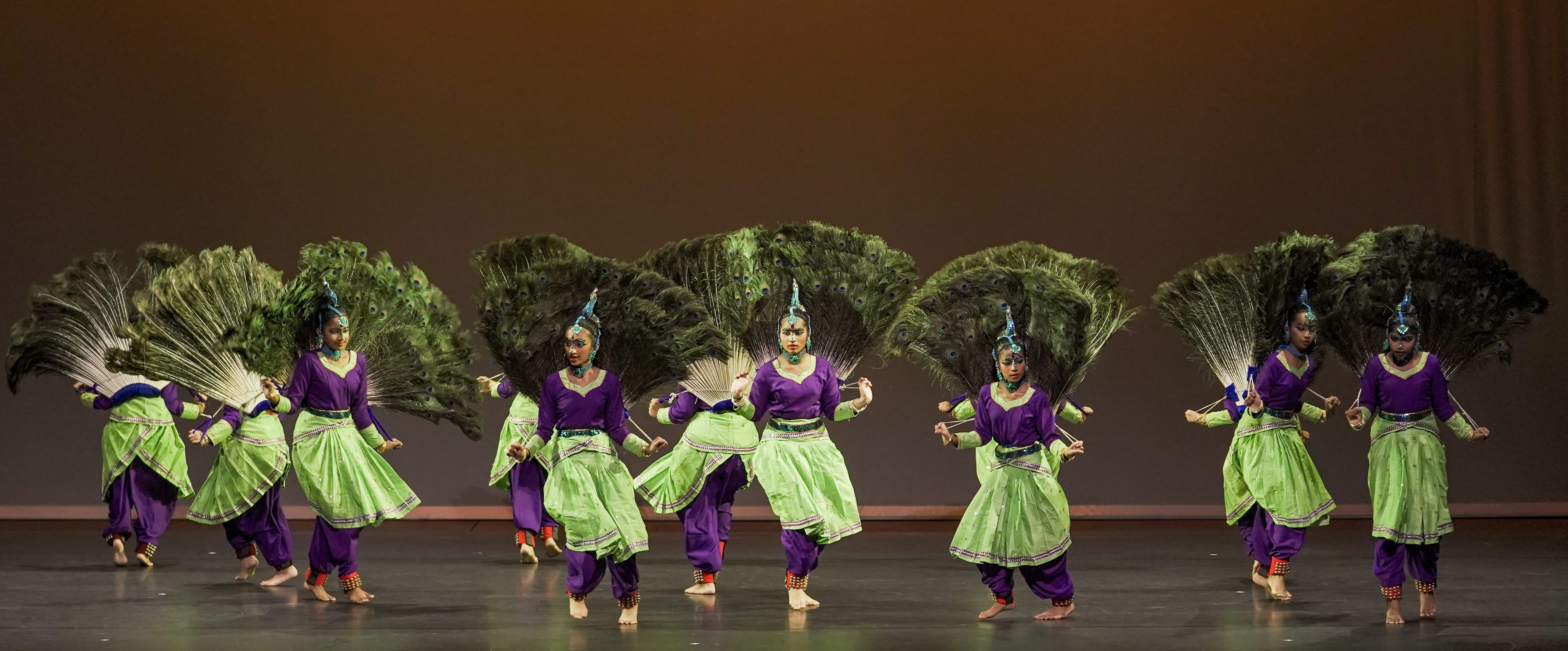 Seven dancers on stage in purple and green costumes holding large peacock feather fans behind their backs.